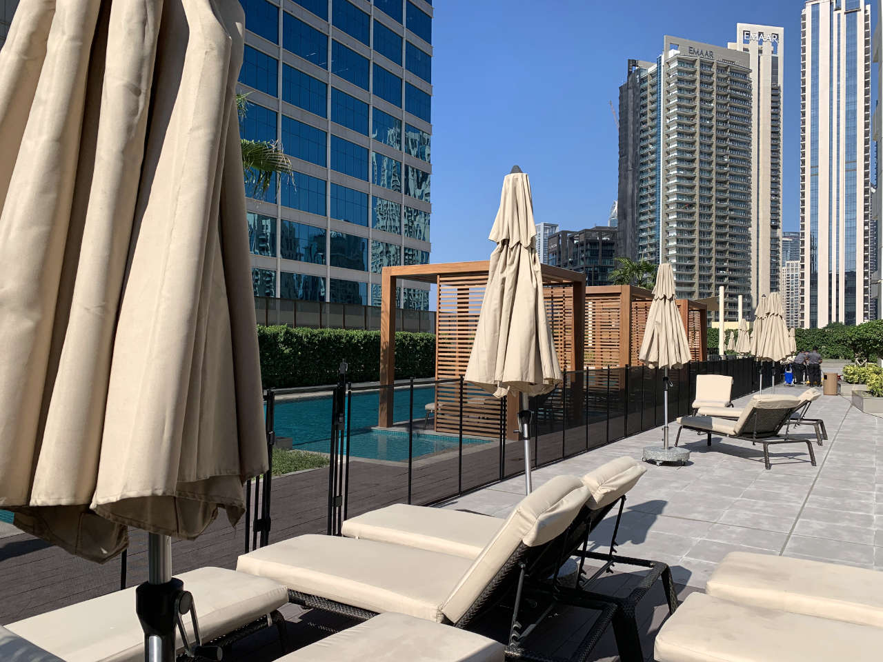 pool-fence-Business-Bay | AMANANET Rooftop pool area with beige umbrellas and lounge chairs, modern glass buildings in the background under a clear blue sky