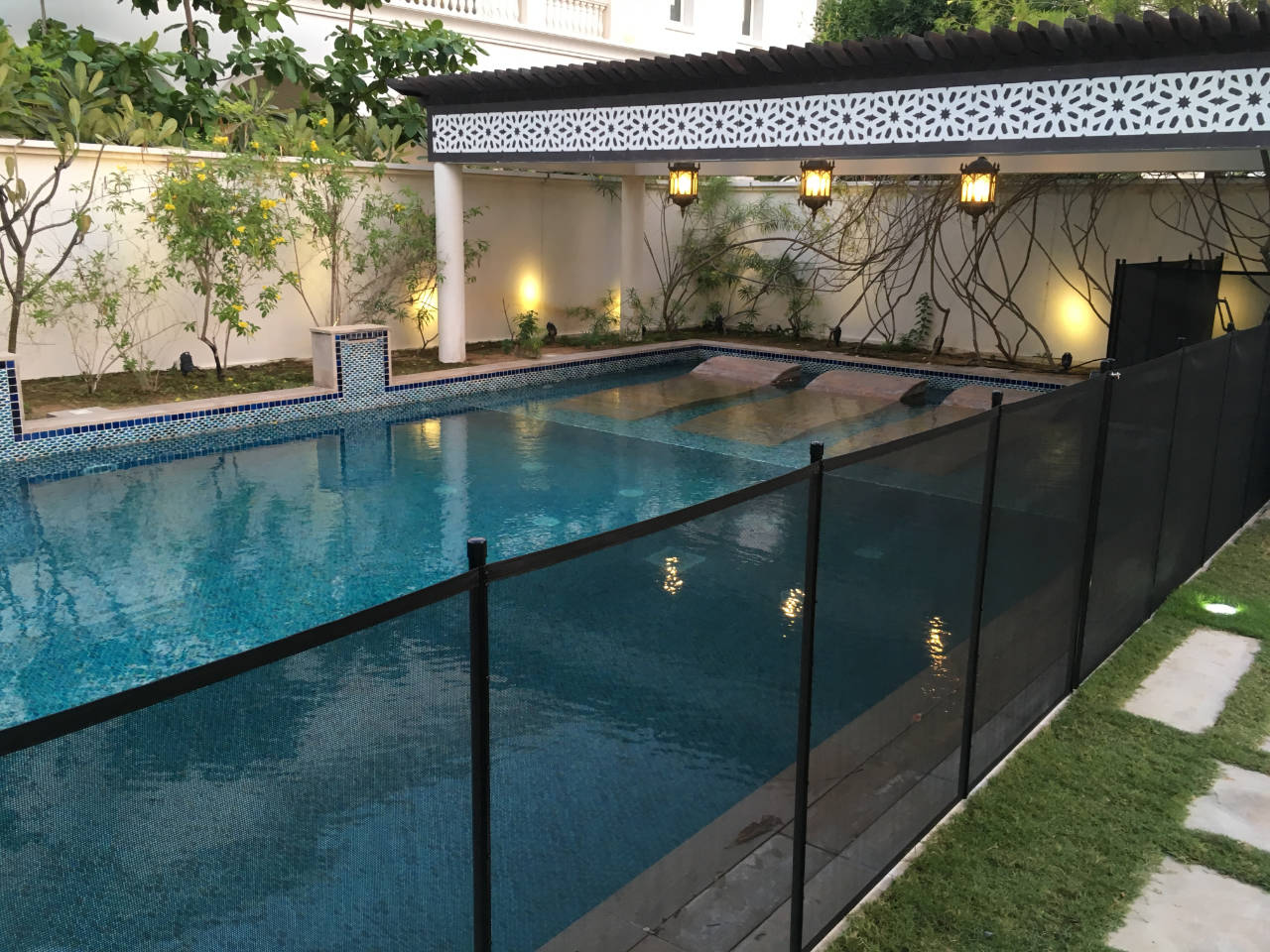 pool-fence-Emirates-Hills | AMANANET Rectangular backyard pool with blue mosaic tiles, glass panel fencing, and a covered lounge area with hanging lanterns.