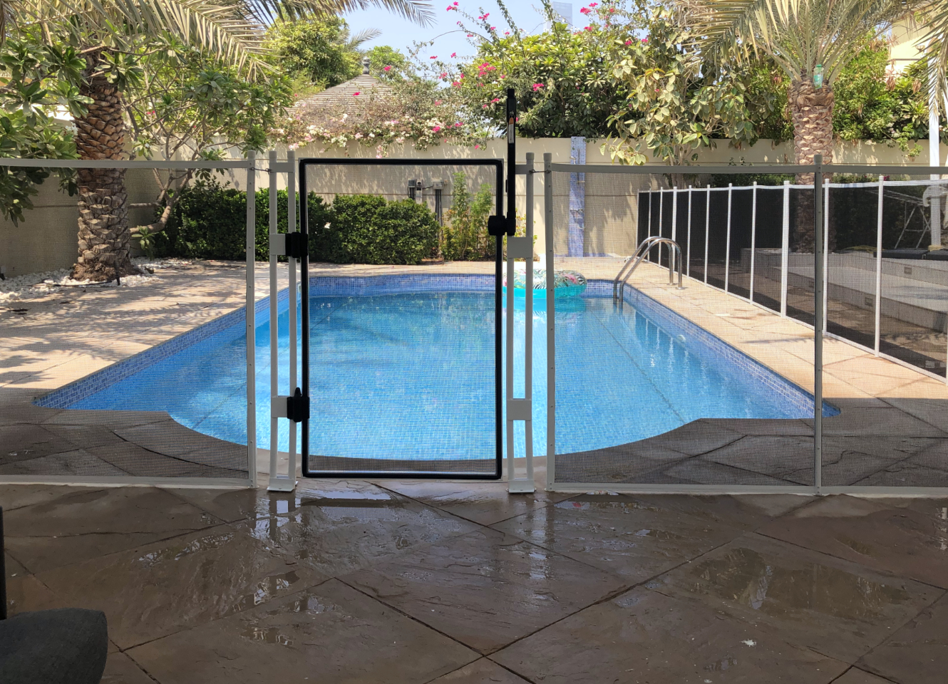 pool-fence-Jumeirah-Park-3 | AMANANET Family pool with a white safety fence and gate enclosing a bright blue rectangular pool; tropical plants and palm trees in the background under a sunny sky.