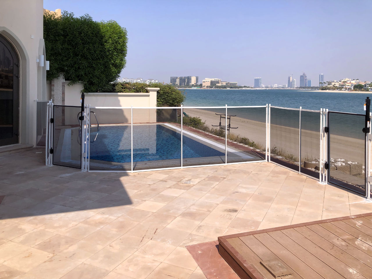 Pool area with glass safety fence overlooking sandy beach and calm sea; city skyline in distance.