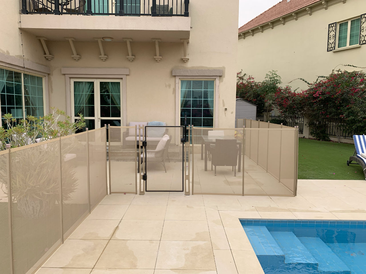 pool-fence-question-3 | AMANANET Backyard patio with beige glass pool fence and gate surrounding seating area beside a rectangular blue pool edge.
